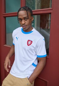 Balts sporta krekls ar ziliem akcentiem, ar sarkano emblēmu, apaļu kakla izgriezumu un īsām piedurknēm. Valkāts kopā ar khaki krāsas šortiem.