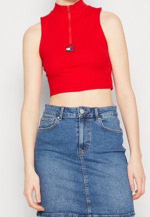 Vrouw draagt een rood, mouwloos cropped top met rits en logo, gecombineerd met een hoge taille blauwe denim rok.