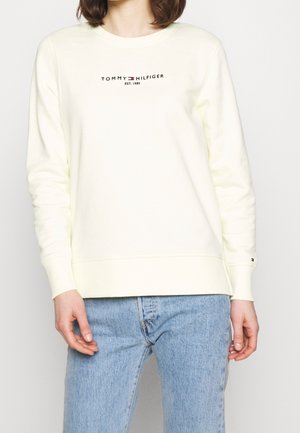 Personne portant un sweat-shirt crème à manches longues Tommy Hilfiger avec logo et un jean bleu clair, debout devant un fond uni.