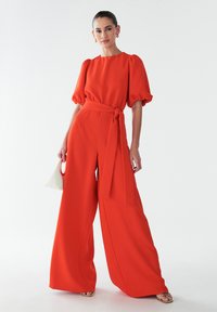 Punane lai jalgadega kombinesoon, millel on kruvitud vöökoht ja puffõlgadega. Sile kangas. Mudel hoiab valget käsitaskut, jalas avatud varvastega kuldsed sandaalid.