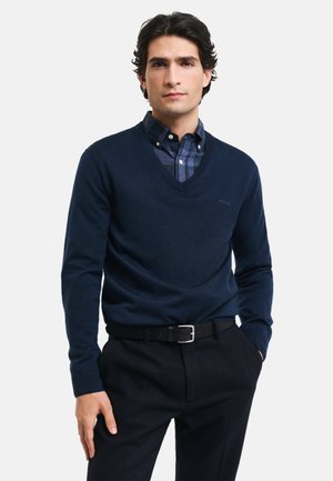 Navy blue v-neck sweater made from soft material, layered over a checkered shirt. Model wears dark trousers and a black belt, with a plain background.