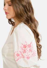 Balta krekla ar pūdētām piedurknēm un rozā ziedu izšuvumu uz kreisās piedurknes. Redzama zelta ķēdes kaklarota. Mīksta tekstūra.