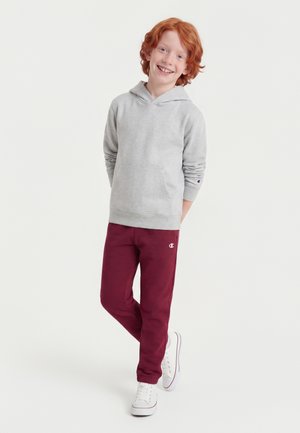 Rødhåret barn smilende, iført en grå hættetrøje, burgunder sweatpants og hvide sneakers, står med hænderne bag ryggen på hvid baggrund.