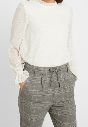 Mujer con blusa blanca texturizada de manga larga con puños fruncidos y pantalones de cuadros grises de cintura alta con cinturón anudado y manos en los bolsillos.