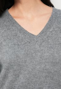 Κοντινό πλάνο ενός ατόμου που φοράει γκρι πλεκτό πουλόβερ με V-λαιμόκοψη, δείχνοντας το λαιμό και την πάνω περιοχή του στήθους.