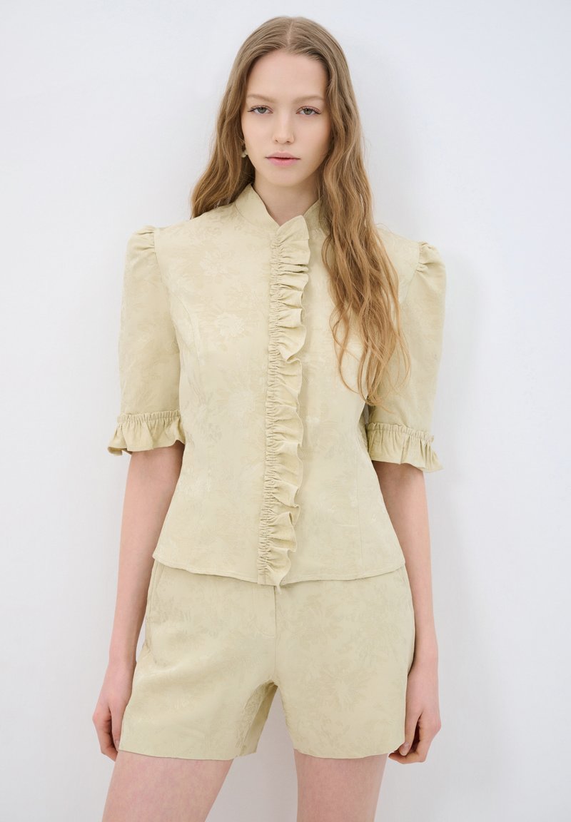 Blusa beige chiaro con maniche corte a sbuffo, davanti arricciato, motivo floreale e colletto alto, abbinata a pantaloni corti coordinati.