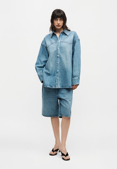 Γυναίκα που φοράει υπερμεγέθη ανοιχτό μπλε τζιν πουκάμισο και τζιν σορτς μήκους μέχρι το γόνατο με μαύρα σανδάλια με τακούνι, στέκεται μπροστά σε ένα απλό φόντο.