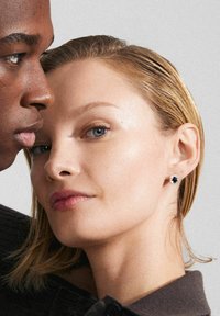 Schwarze Steckerohrringe mit silbernen Akzenten, die eine runde Form und einen funkelnden Rand haben. Das Model hat glattes, blondes Haar und eine makellose Haut.