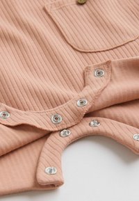 Close-up of peach ribbed fabric with silver snap fasteners along a curved edge and a small pocket with a button at the top.
