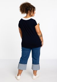 Marineblaue T-Shirt mit weißen Akzenten an den Schultern, kombiniert mit cropped Jeanshosen mit einem hellblauen Saum. Dazu werden weiße Sneakers getragen.