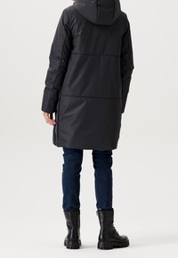 Abrigo largo negro acolchado con capucha, que presenta cremalleras laterales y una costura en la cintura, combinado con jeans oscuros y botas de combate negras.