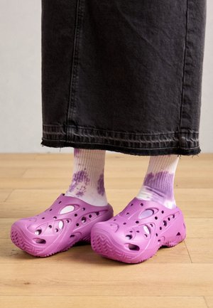 Fioletowe gumowe chodaki z otworami wentylacyjnymi noszone z białymi i fioletowymi skarpetkami tie-dye oraz długą czarną spódnicą, stojące na drewnianej podłodze.