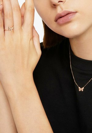 Woman wearing matching rose gold butterfly ring and necklace, with hand near face and neutral lip expression, black top visible.
