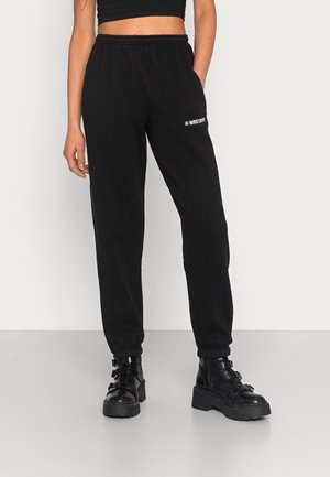 Vrouw draagt zwarte high-waisted joggers met de tekst "WRSTBHVR" op de dij, zwarte crop top en zwarte chunky buckle boots op een grijze vloer.