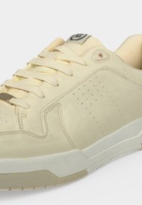 Baskets couleur crème avec tige en daim, panneau latéral perforé, fermeture à lacets et semelle extérieure blanche en caoutchouc texturée.