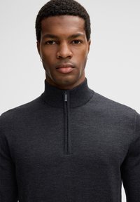Dunkelgrauer Zip-Kragen-Pullover aus glattem Strickmaterial. Besitzt einen hohen Kragen und einen halben Reißverschluss sowie eine taillierte Silhouette.