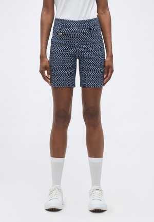 Osoba nosiaca námorníckym modrým vzorovaným šortkám, biele ponožky crew, biele tenisky a bielu košeľu vpustenú dnu, stojaca pred jednofarebným pozadím.