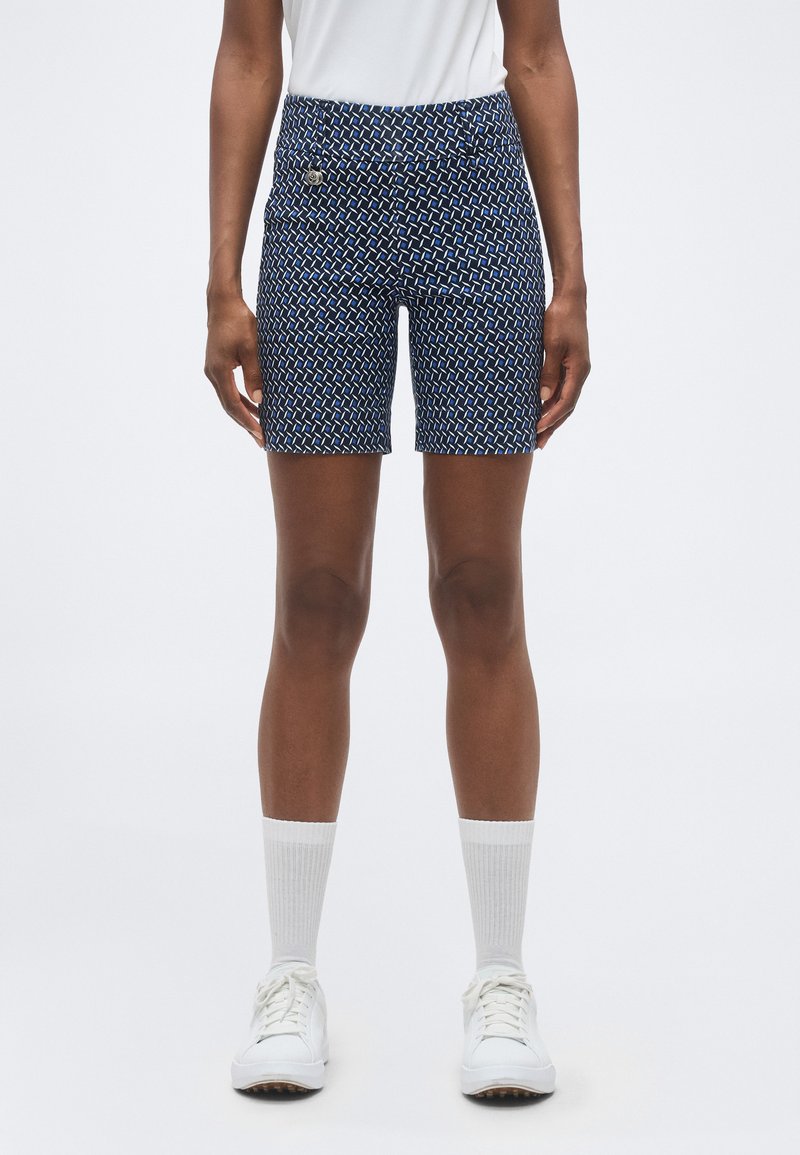 Άτομο που φοράει μπλε σκούρο σορτς με σχέδια, λευκές κάλτσες τύπου crew, λευκά αθλητικά παπούτσια και λευκό πουκάμισο σεμνά μέσα στο παντελόνι, στέκεται μπροστά σε μονόχρωμο φόντο.