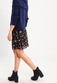 Pull en laine bleu marine avec col roulé, jupe noire à fleurs, et bottines à talons épais avec panneaux élastiques, mettant en valeur des textures superposées.