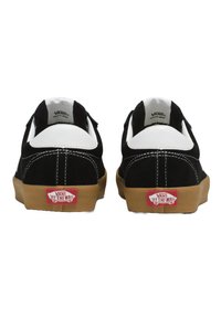 Fekete velúr sneakerek fehér bőr részletekkel, gumi talppal és piros "Vans Off the Wall" logóval a saroknál. Kétvarrás látható.
