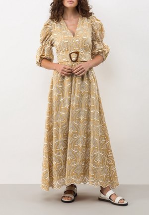 Kobieta w beżowej sukience maxi z wzorem, z bufkami na rękawach, paskiem w talii i białych sandałach, stojąca na jednolitym tle.