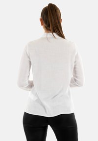 Blusa blanca de manga larga con tejido texturizado, cuello clásico, dobladillo curvado y puños abotonados. Combinada con pantalones negros ajustados.