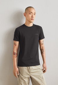 T-shirt nera in cotone con maniche corte e scollo rotondo, caratterizzata da un piccolo logo ricamato sul petto. Indossata con pantaloni cargo beige chiaro.