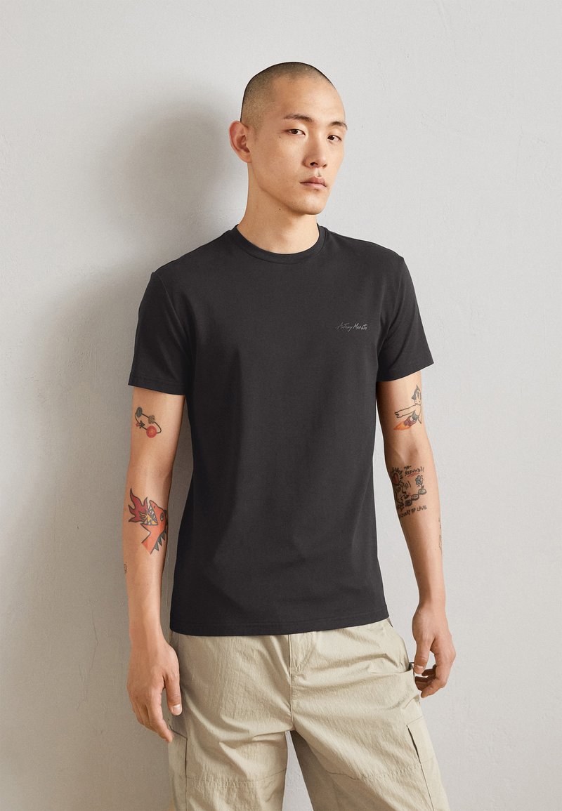 T-shirt nera in cotone con maniche corte e scollo rotondo, caratterizzata da un piccolo logo ricamato sul petto. Indossata con pantaloni cargo beige chiaro.