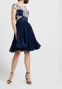 Marinblå klänning med utsvängd kjol, med en djup V-ringning och spetsappliké-design på överdelen. Försedd med korta, utsmyckade ärmar.