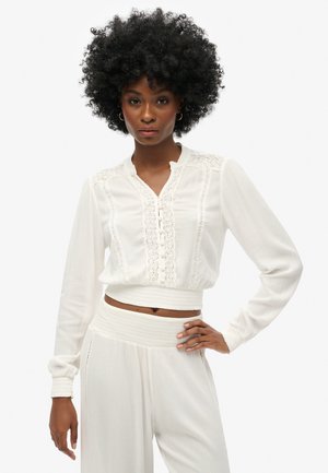 Blusa blanca, corta, con mangas largas, detalles de encaje y cierre de botones. Tela ligera con una textura suave y cintura ajustada.
