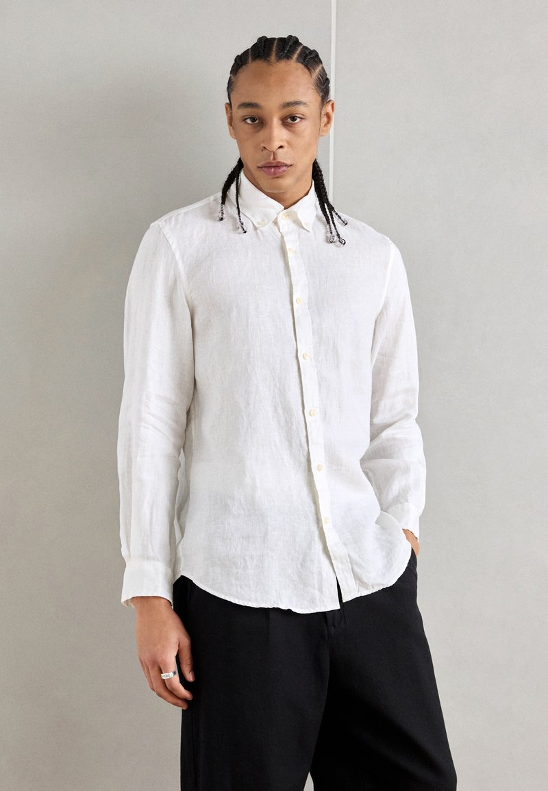 Chemise en lin blanche à manches longues, texture subtile et ourlet incurvé, associée à un pantalon noir ample. Un homme se tient les mains dans les poches.