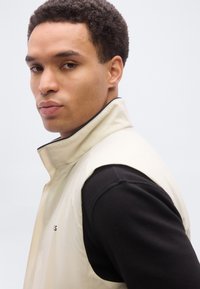 Νεαρός άνδρας με κοντά σγουρά μαλλιά, φοράει μπεζ γιλέκο χωρίς μανίκια πάνω από μαύρο πουλόβερ με μακριά μανίκια, κοιτάζει πάνω από τον ώμο του.