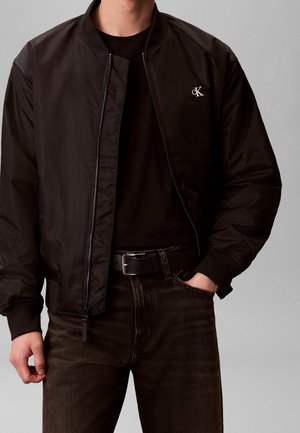 Mand iført sort bomberjakke med Calvin Klein-logo, sort bælte, mørke jeans og sort skjorte, hånd i lommen, mod grå baggrund.