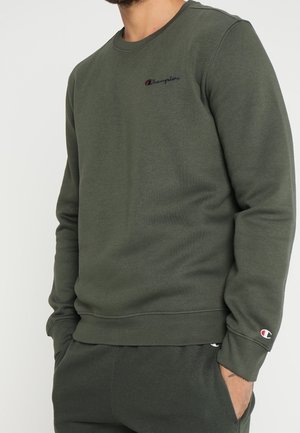 Homme portant un sweat-shirt vert olive à manches longues et un pantalon assorti, avec de petits logos Champion sur la poitrine et le poignet, les mains dans les poches.