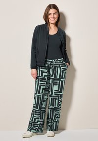 Czarna kurtka bomberka na ciemnym topie na ramiączkach, w połączeniu z szerokimi spodniami w miętowym kolorze, ozdobionymi wyrazistym czarnym geometrycznym wzorem. Białe sneakersy.