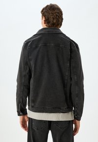 Chaqueta de denim negra con un acabado ligeramente desgastado, cuello clásico y puños abotonados. Presenta una silueta recta y sutiles detalles de costura.