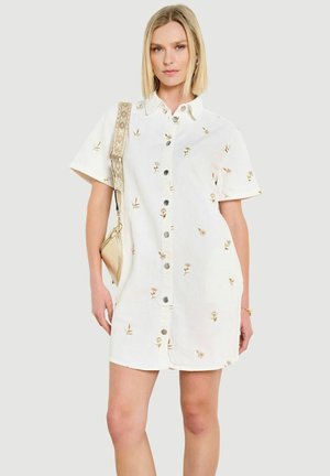 Kobieta w białej sukience koszulowej z małymi kwiatowymi nadrukami, nosząca złotą torebkę na ramieniu z wzorzystym paskiem.