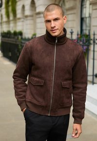 Mann mit kurzen Haaren, der eine braune Wildlederjacke mit Reißverschluss und Taschen trägt, steht draußen auf einer Stadtstraße.