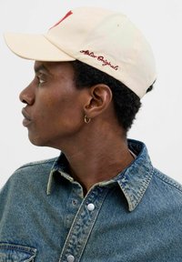 Beige cap with red embroidery reading “Atelier Originals.” Worn with a blue denim shirt. Profile view highlighting the cap's curved brim.