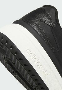 Zapatilla negra con parte superior de cuero texturizado, paneles laterales elásticos y una entresuela blanca con una suela exterior de goma negra, detalle de logotipo en relieve.