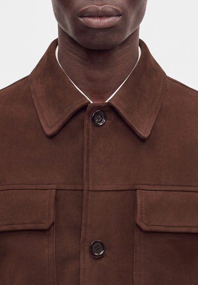 Gros plan d'une personne portant une veste en daim marron avec un grand col, deux poches poitrine et des boutons noirs attachés en haut.