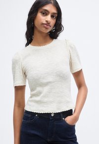 Τοπ από κρεμ πλεκτό ύφασμα με κοντά puff μανίκια και ραμμένη άκρη με κυματιστό σχέδιο. Συνδυάζεται με σκούρο τζιν παντελόνι.