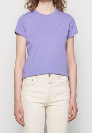 Femme portant un t-shirt lilas ajusté à manches courtes avec un petit logo turquoise sur la poitrine et un jean taille haute blanc cassé.