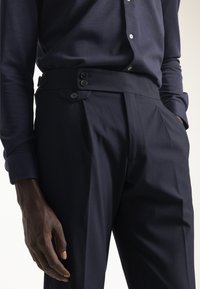 Pantalones de corte entallado en azul marino con una cinturilla alta, que cuentan con dos cierres de botón y pliegues marcados para un diseño estructurado. Textura de tejido suave.