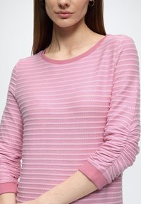 Lichtroze top met lange mouwen, met getextureerde strepen en geplooide details bij de mouwen. Voorzien van een ronde halslijn en een zachte stofafwerking.