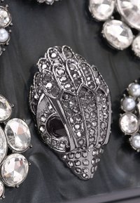 Zwarte metalen broche met een ingewikkeld bladachtig ontwerp, versierd met verschillende facetten van heldere en parelachtige stenen, geplaatst tegen een donkere achtergrond.