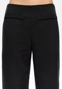 Pantalones negros de cintura alta con textura y bolsillos con ribete horizontal en la parte delantera, mostrados desde la cintura hasta la parte superior del muslo.