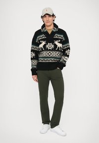 Juodas megztinis su baltais elniais ir geometriniais raštais; apykaklės iškirptė. Alyvuogių žalios kelnės ir baltos sportbačiai papildo aprangą.