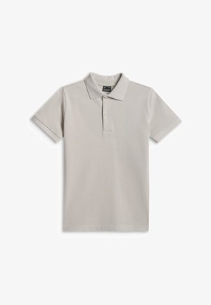 Garçon debout devant un fond gris, portant un polo beige, un pantalon cargo sombre et des baskets blanc-gris, regardant devant avec une expression neutre.