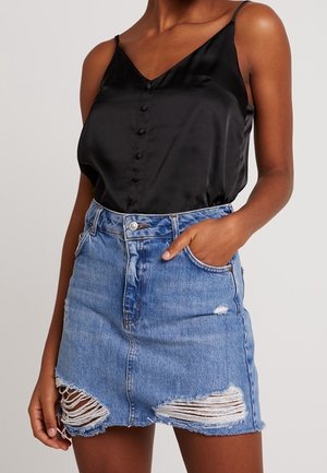 Vrouw draagt een zwart satijnen camisole met knopen en een versleten blauwe denim minirok, met haar hand in de zak.